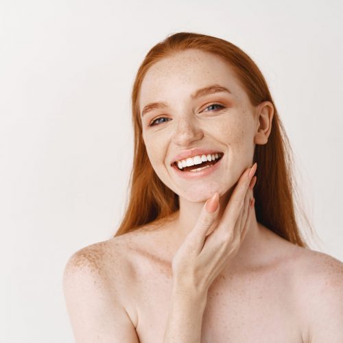Beauty. Close-up of young beautiful redhead woman smiling at camera, touching perfect clean skin on face and looking happy, standing naked over white background.